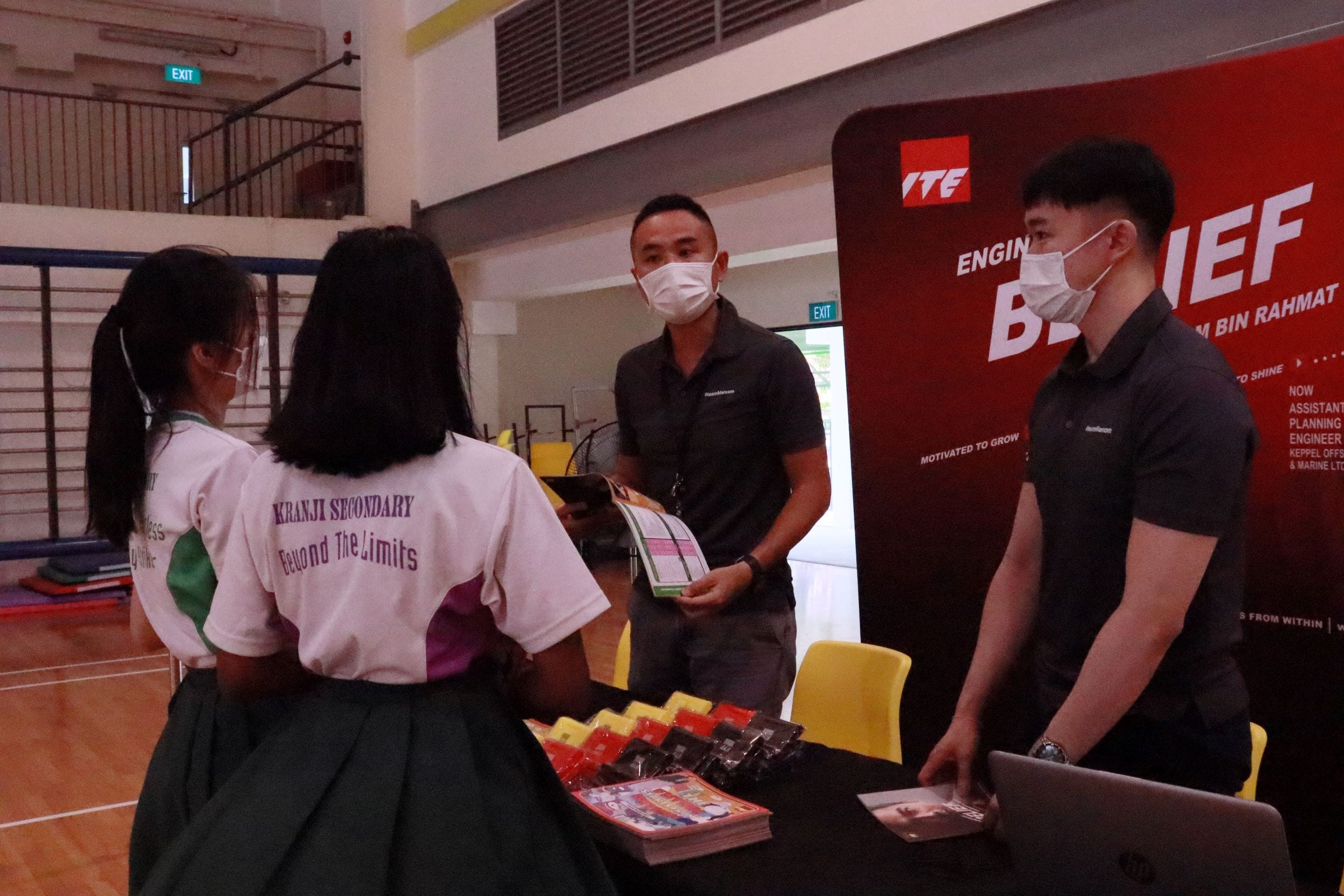 Two Kranji Secondary students speak with masked ITE representatives at a booth inside a school hall.